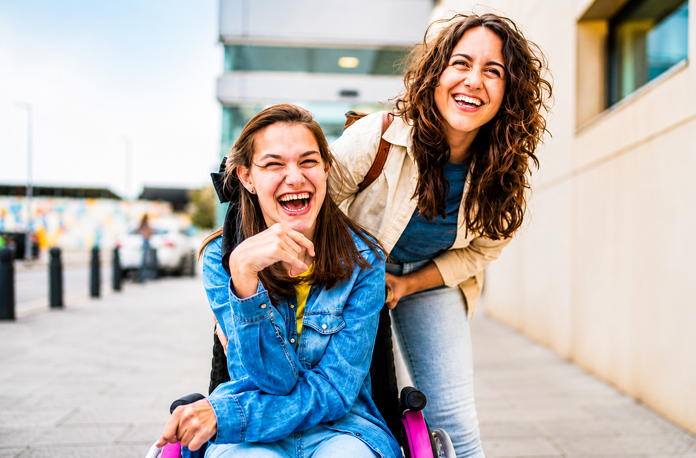 Two smiling people outdoors, one in a wheelchair, posing beside a building.