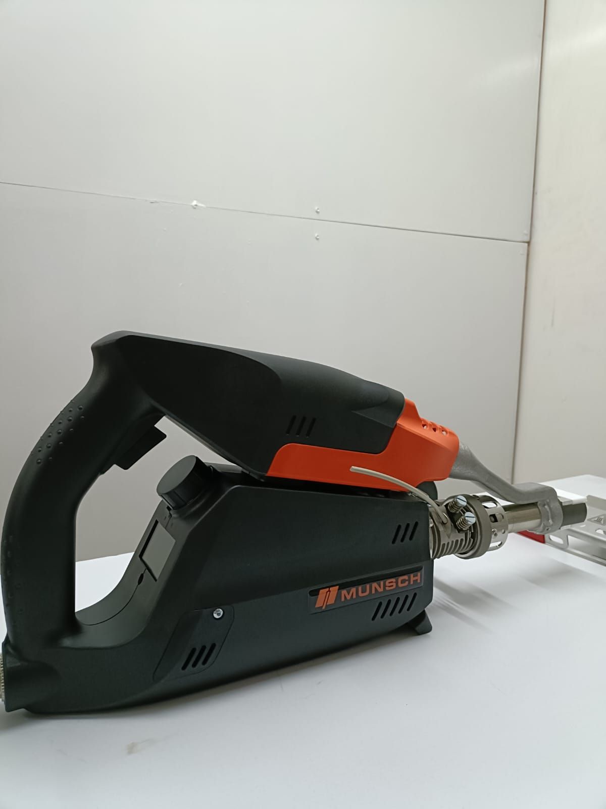 Herramienta eléctrica de mano de color negro y naranja sobre una superficie blanca, con un accesorio plateado a la derecha.