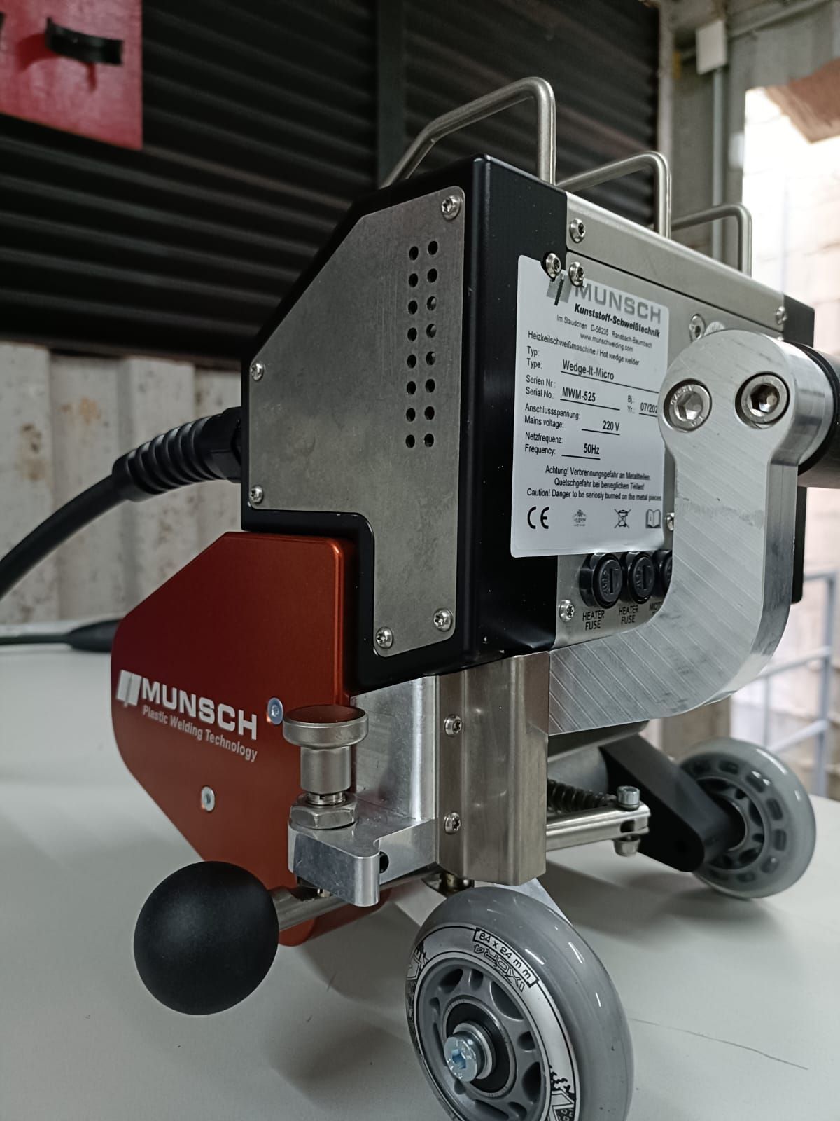 Una máquina de soldar con ruedas, roja y plateada. Tiene cable de alimentación, asa y una bola negra.