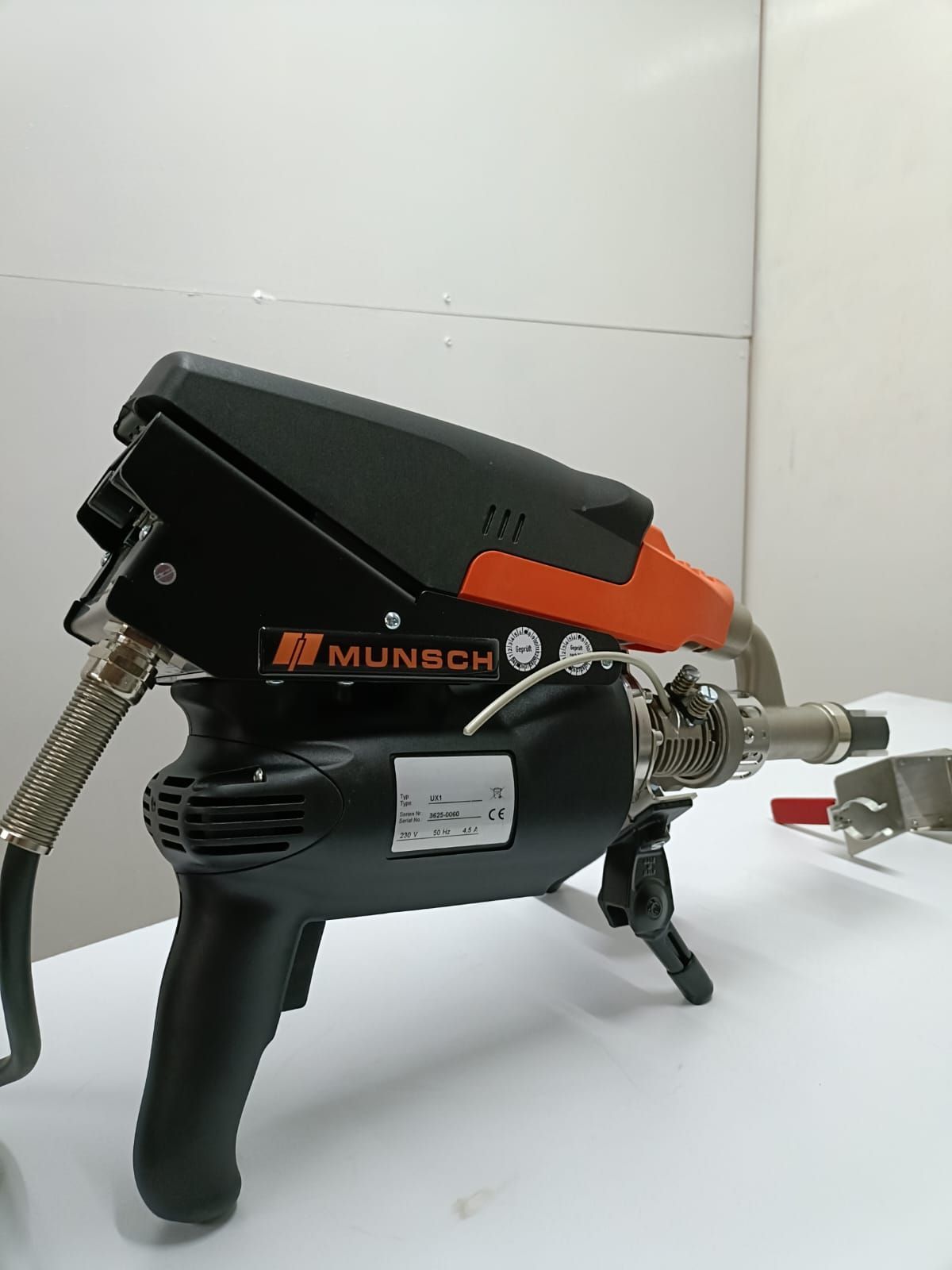 Herramienta industrial negra y naranja con manguera y boquilla sobre un suelo blanco contra una pared de azulejos.
