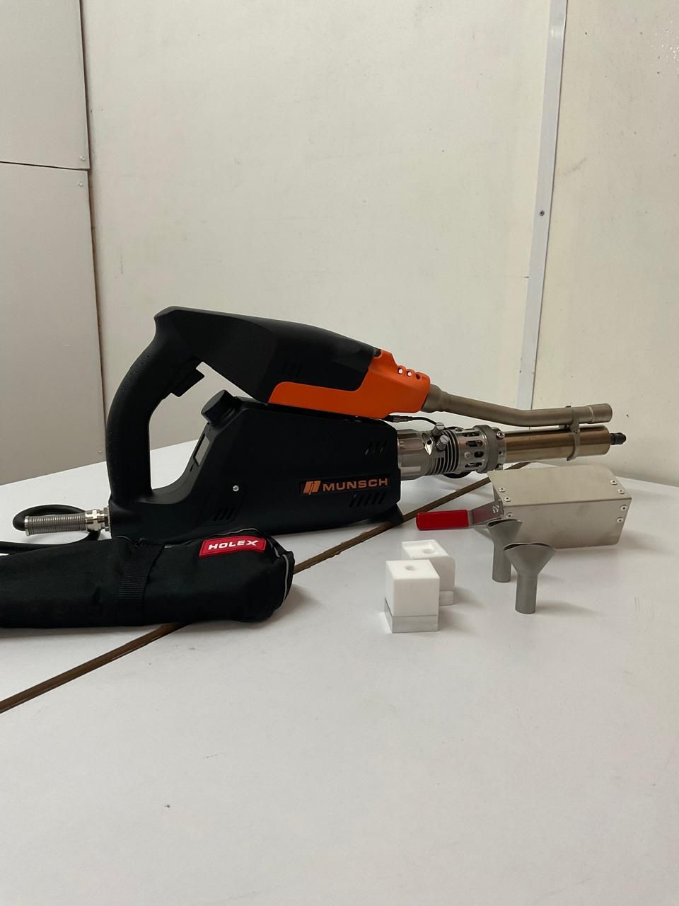 Herramienta eléctrica inalámbrica negra y naranja sobre una mesa con accesorios metálicos y pequeños bloques blancos.