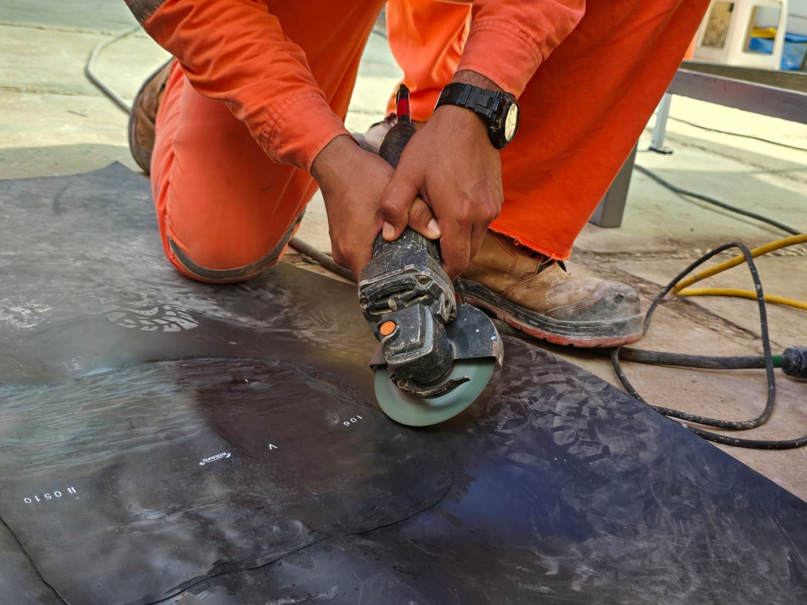 Trabajador con mono naranja usando una amoladora angular sobre una superficie de suelo oscura con cables cerca.