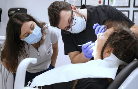 Dentist and assistant examining a patient in a dental chair, both wearing masks and gloves.
