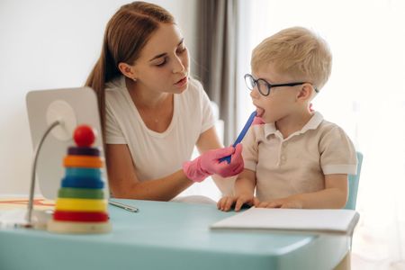 Adult assisting child with a pen at a table, with a colorful stacking toy in the foreground