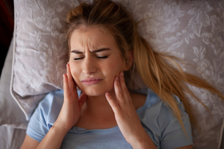 Woman lying in bed, holding her face with eyes closed, appearing to have a headache