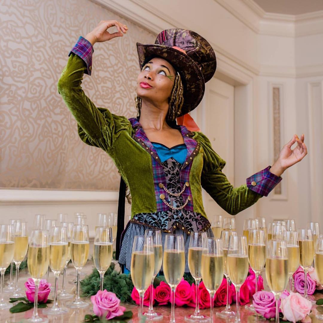 A woman in a mad hatter costume is standing in front of a table full of champagne glasses.