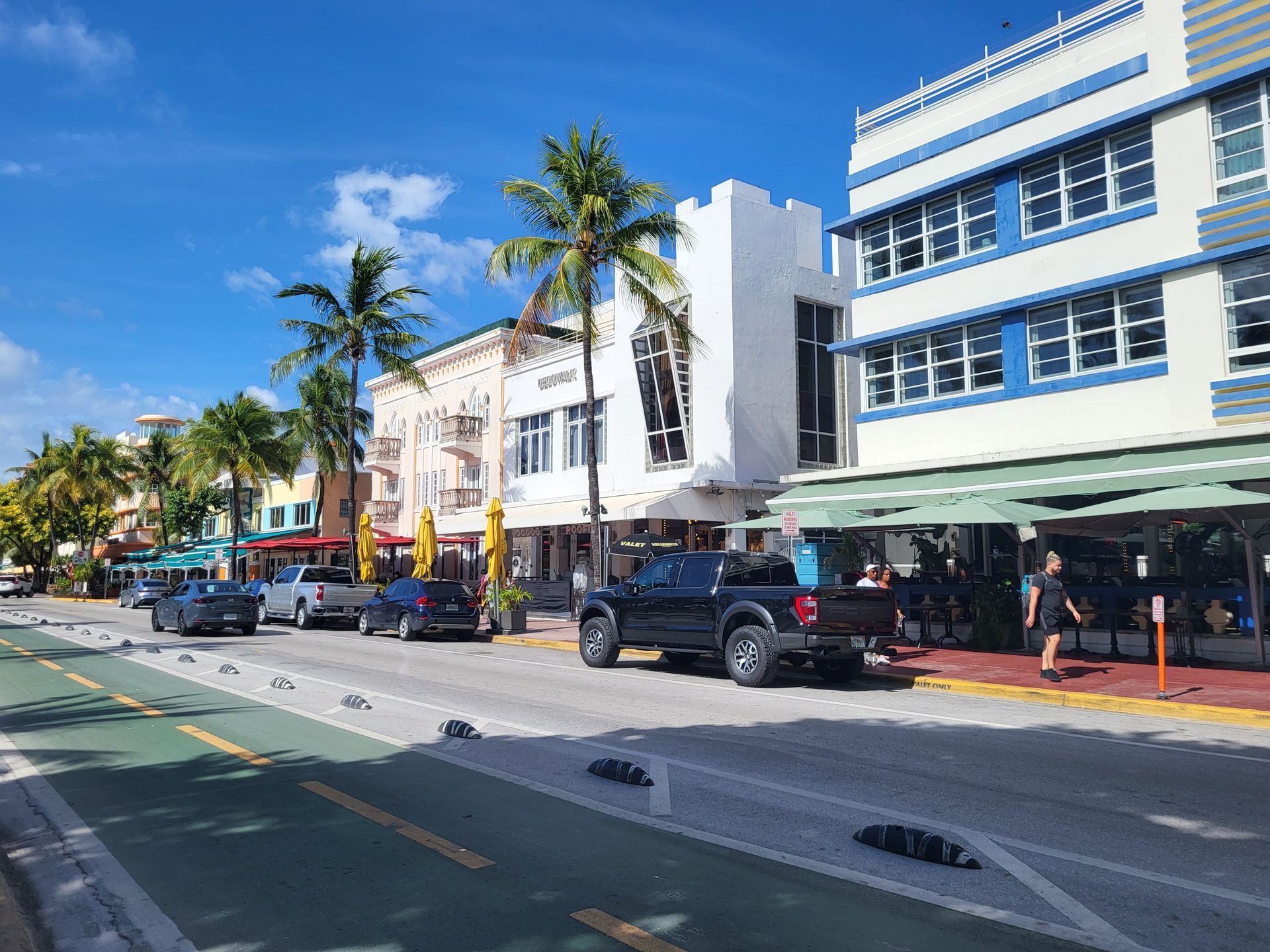 Sunny street with palm trees, parked cars, and white storefront buildings along a blue sky roadway