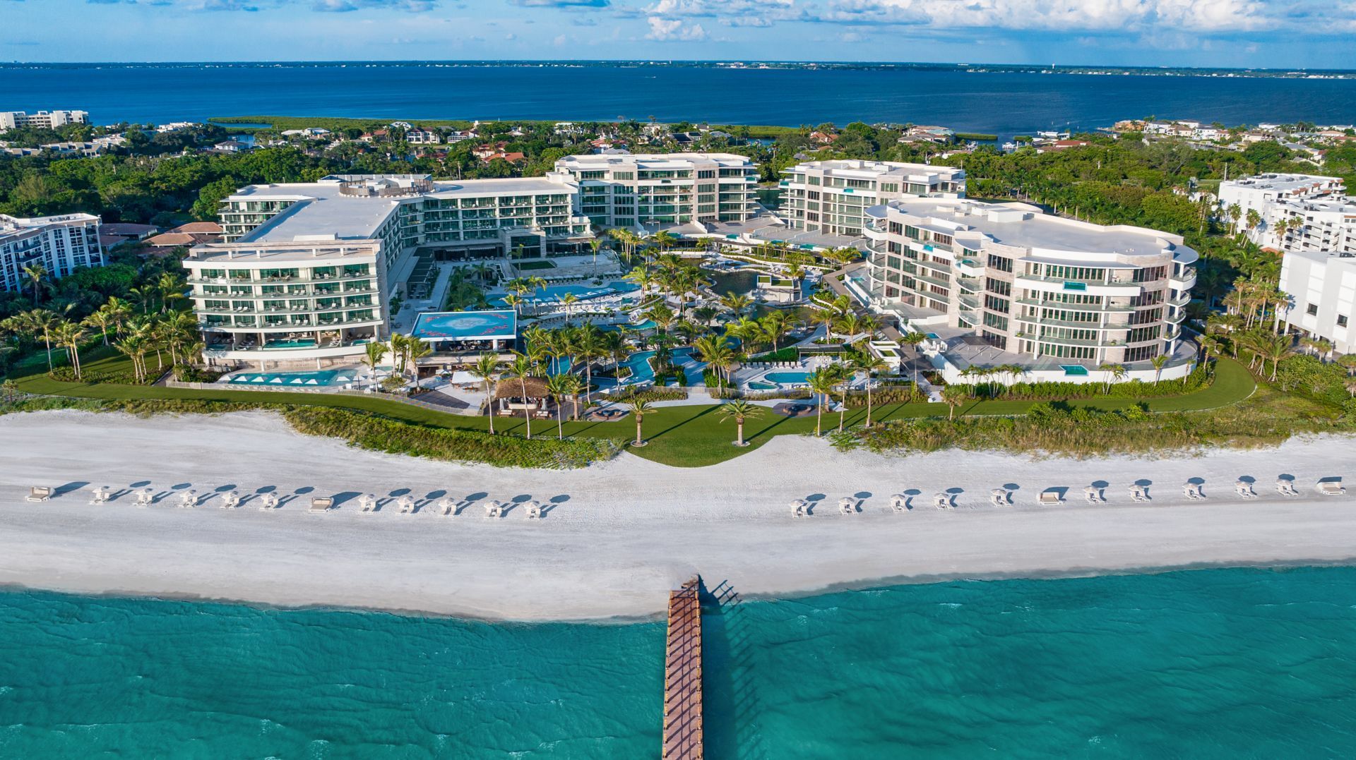 Aerial view of a beachfront resort with pools, white sand beach, and a pier extending into turquoise water