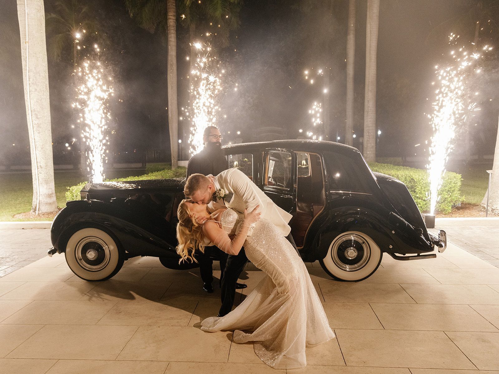 A bride and groom kissing in front of a black car.