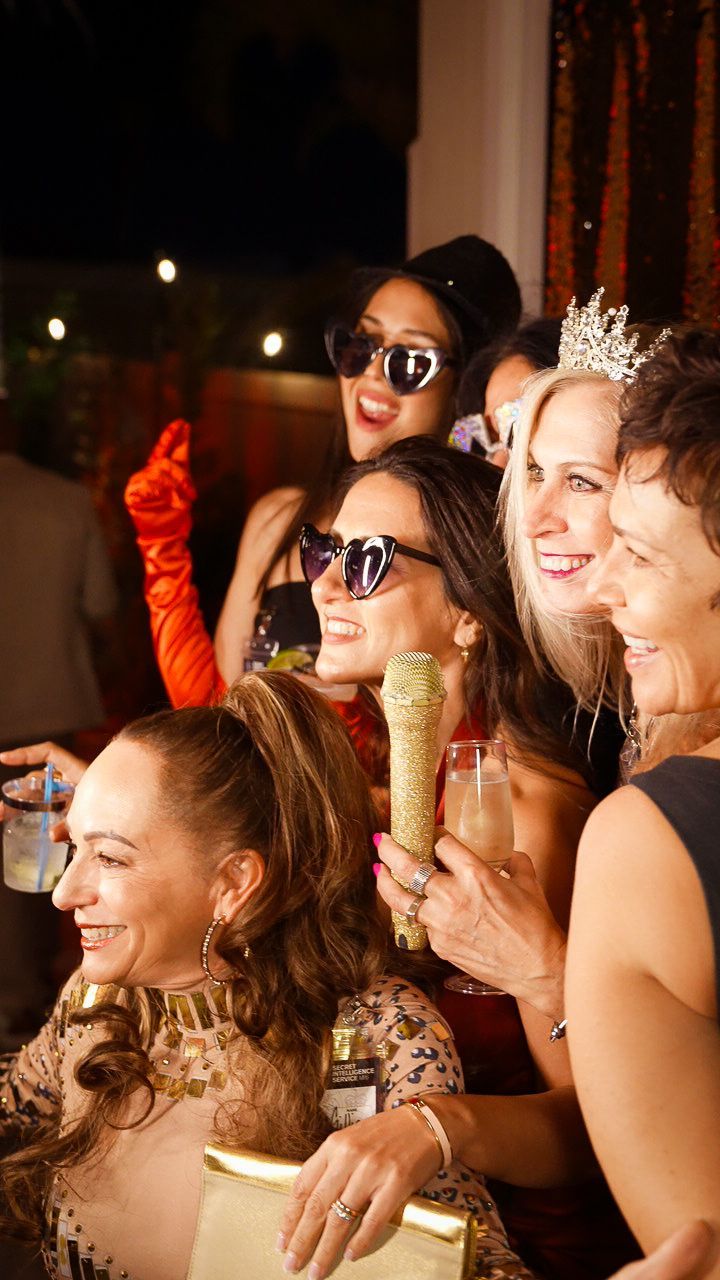 Friends in festive outfits and sunglasses posing together at a lively party, smiling and holding drinks.