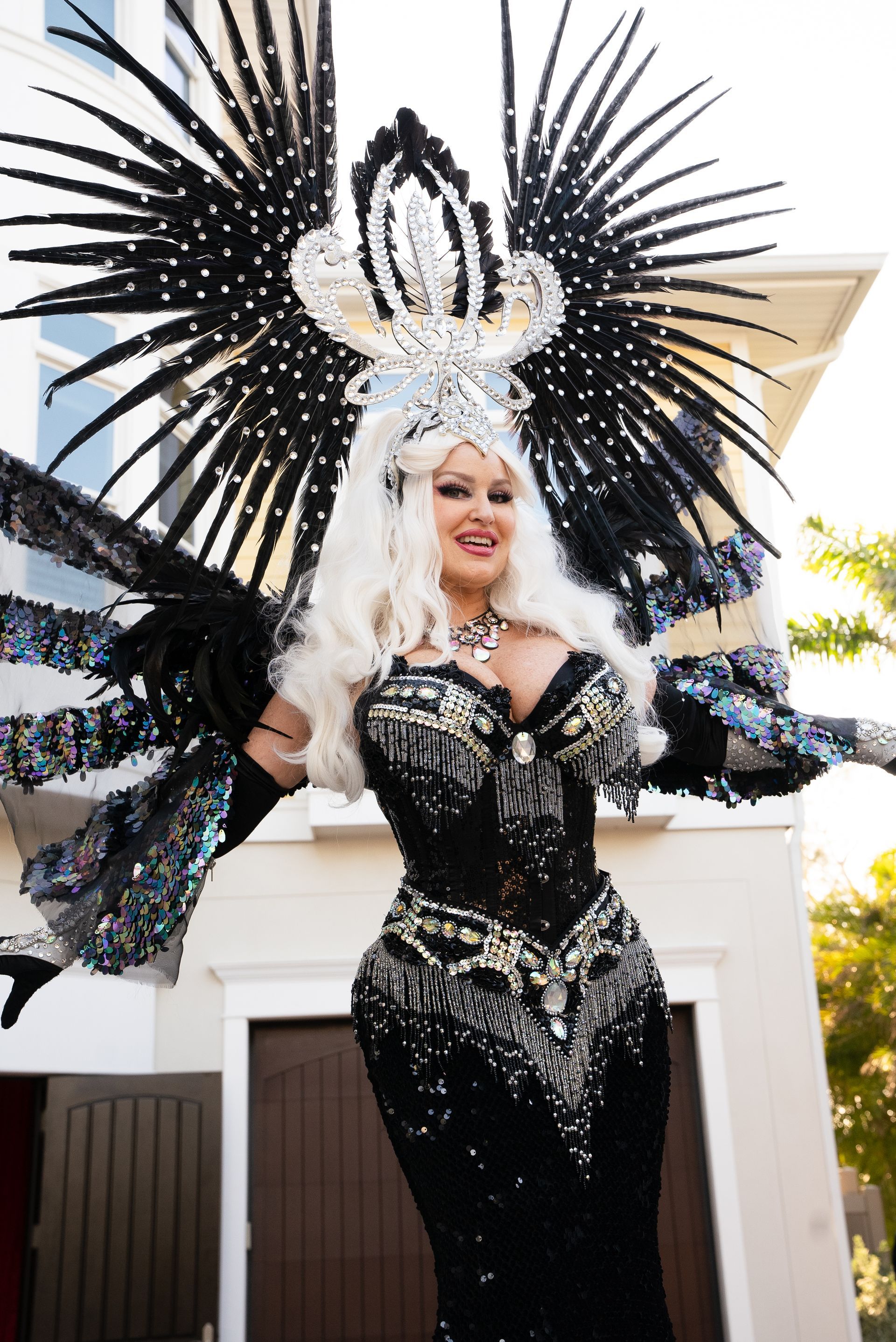 Person in a black sequined costume with a feathered headdress, posing outdoors in front of a house.