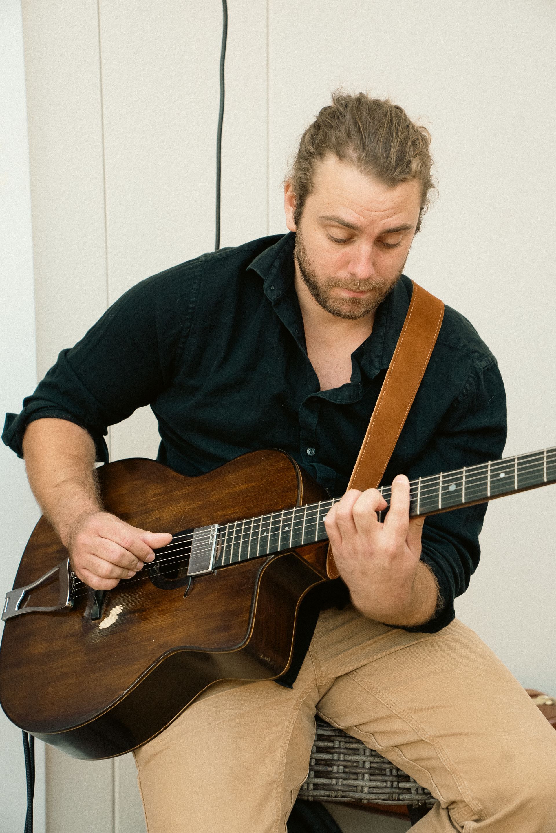 Man seated indoors playing a brown acoustic guitar, wearing a dark shirt and tan pants.