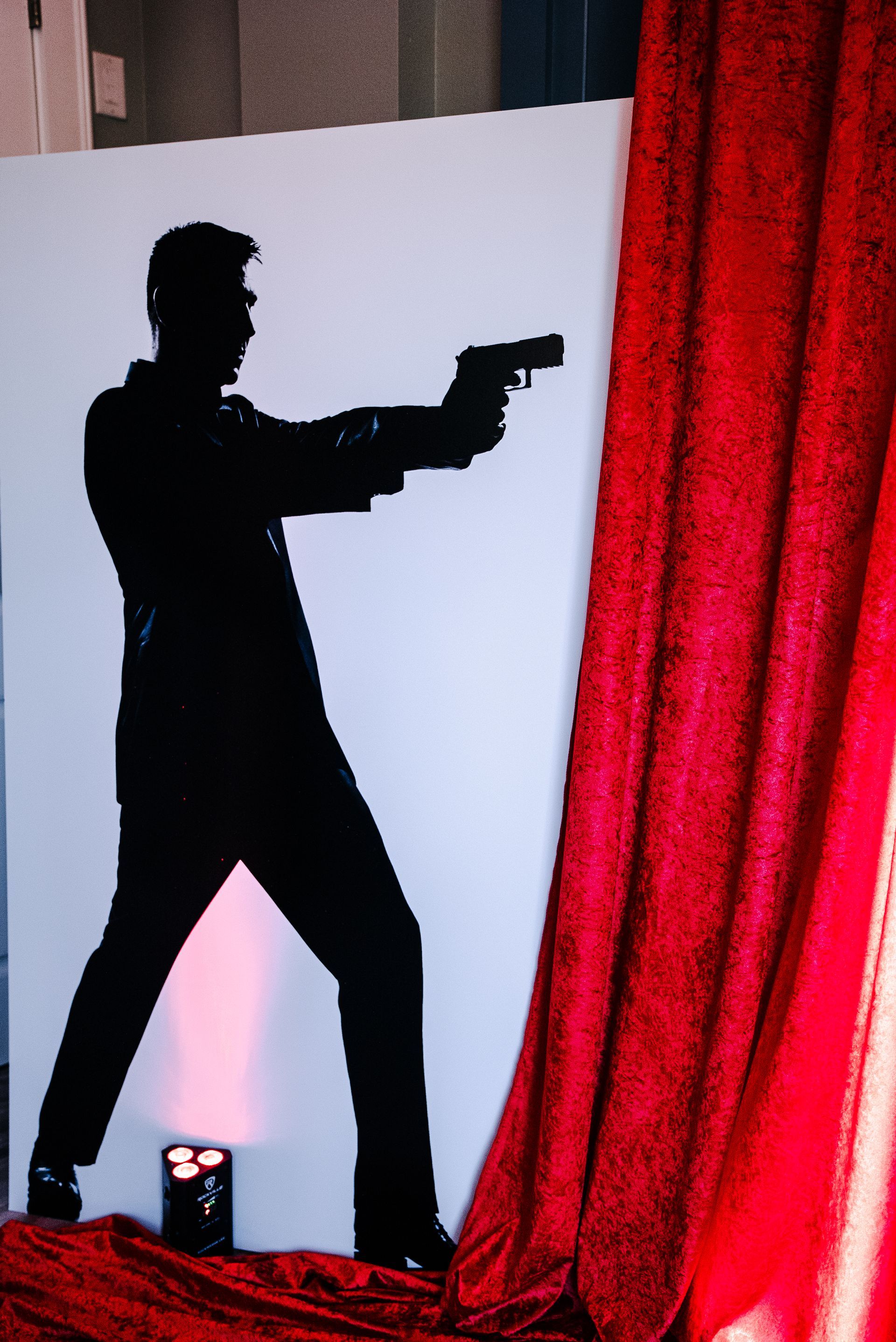 Silhouette of a person aiming a handgun beside a red curtain on a lit stage