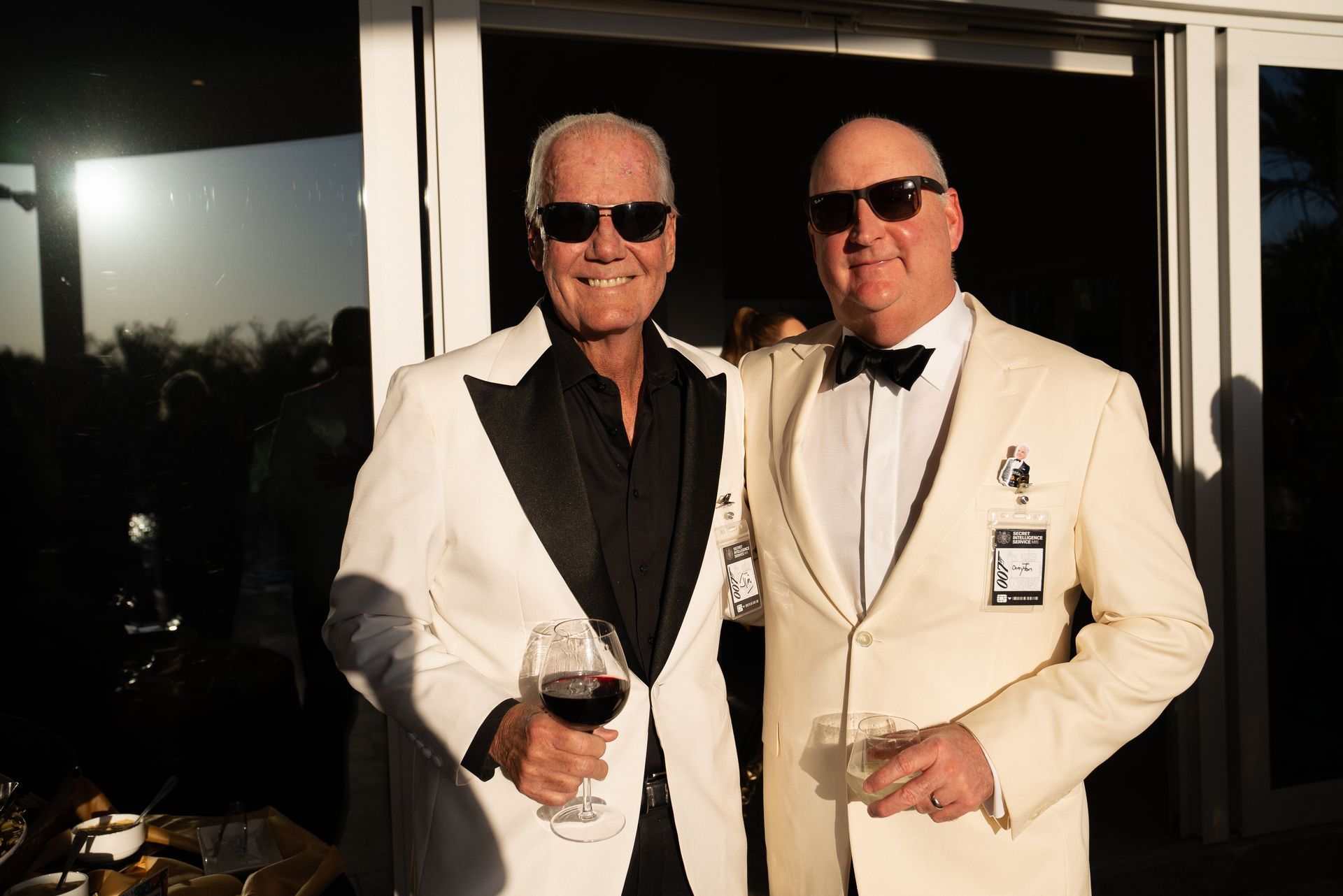 Two men in white tuxedos and sunglasses holding drinks, standing by a sliding glass door.