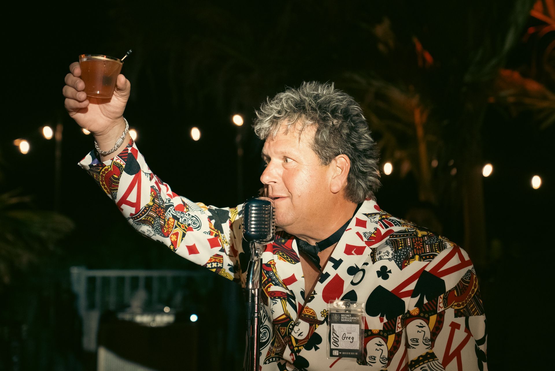 Person raising a drink beside a microphone, wearing a colorful patterned shirt at a nighttime event
