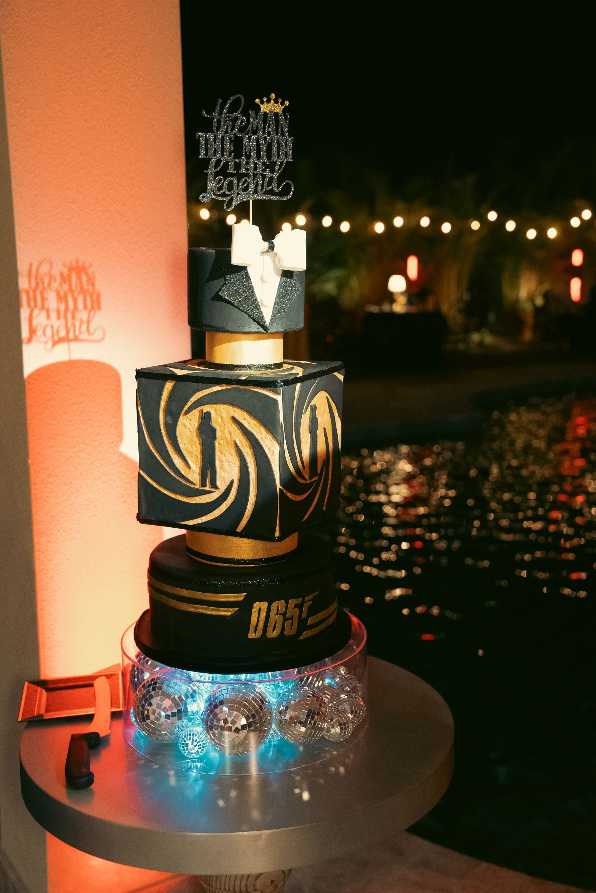 Decorative layered cake on a glass stand at a nighttime waterfront event with lights in the background