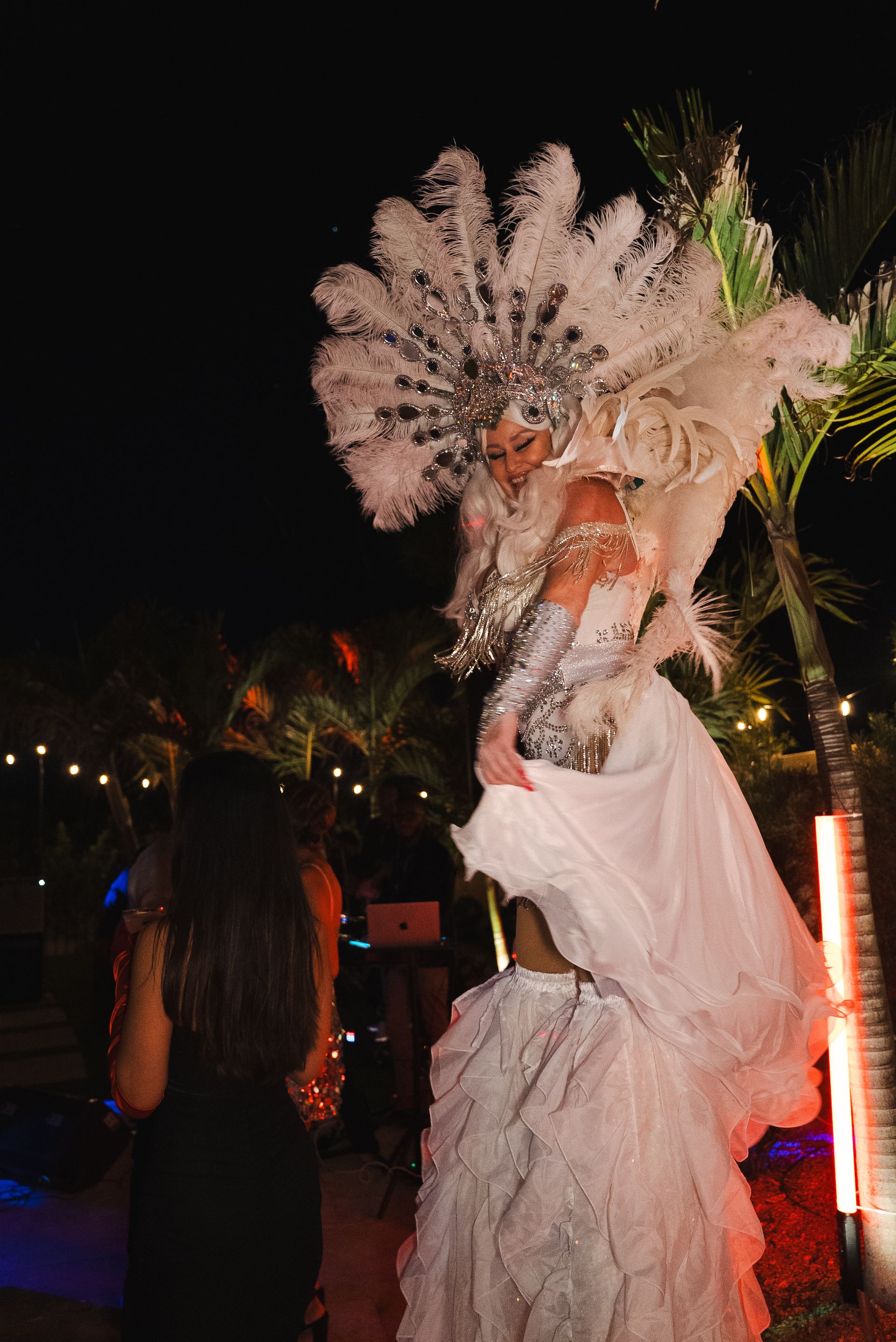 Person in a large white feathered costume posing outdoors at night with palm trees and colorful lights