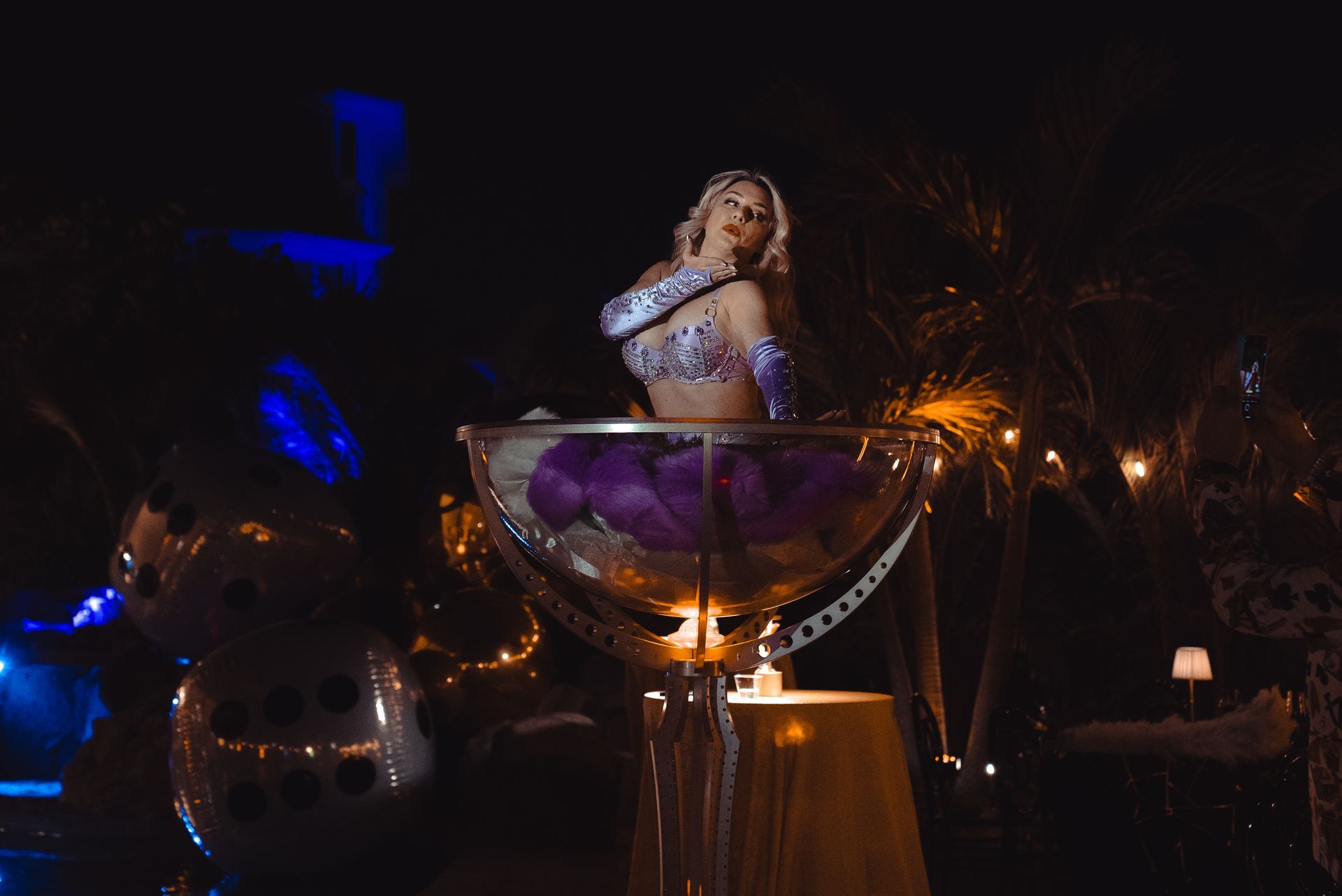 Singer performing onstage inside a giant martini glass, lit by blue and amber lights.