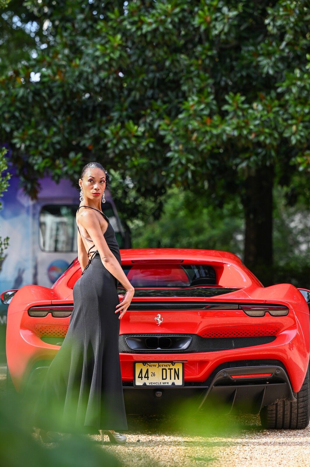 Woman in a black dress standing beside a red sports car in a leafy outdoor setting