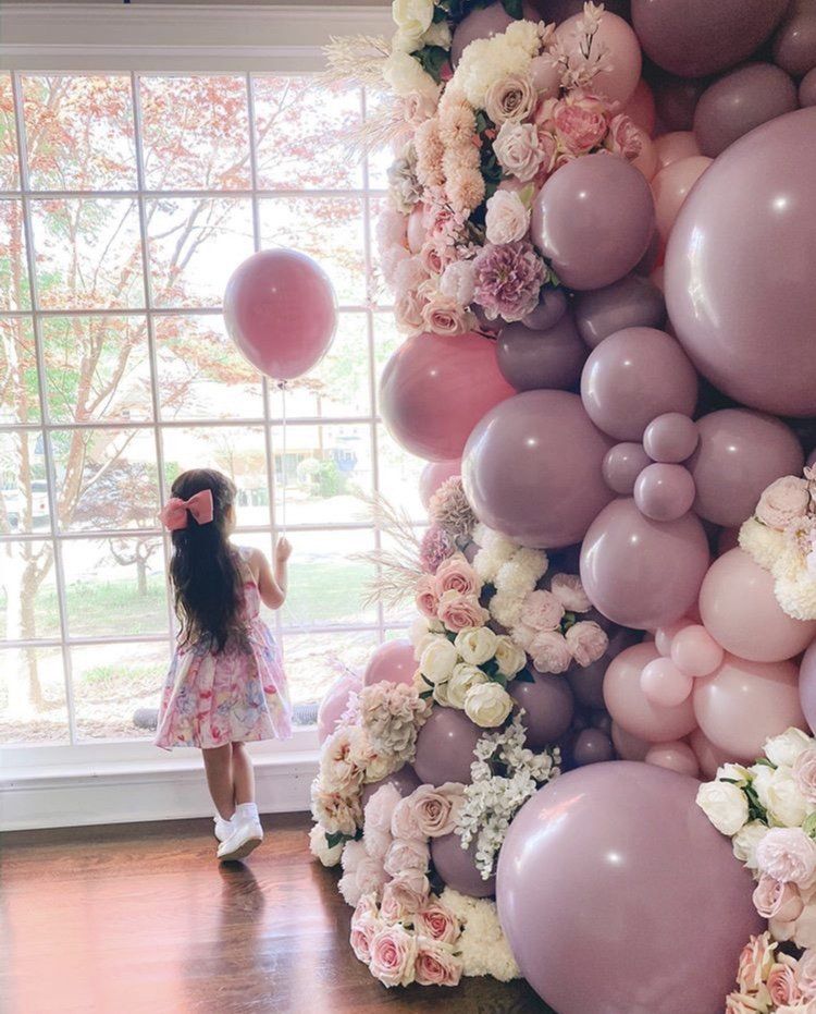 Girl in a pink dress standing by a large pastel balloon and flower wall in a bright room