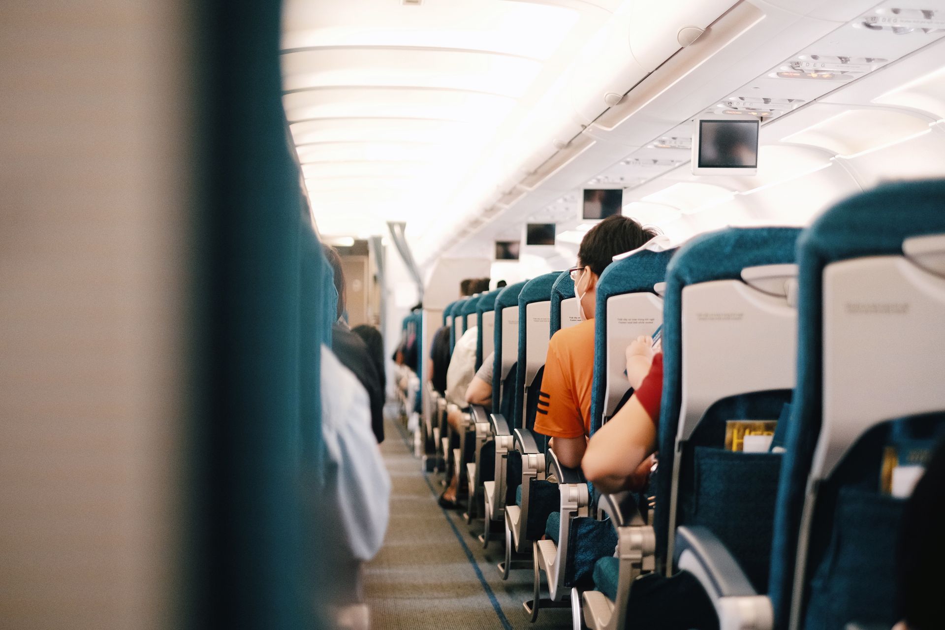 Passengers seated inside an airplane cabin, with blue seats and bright overhead lighting.