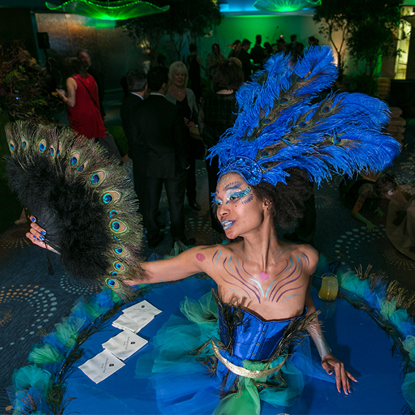 Costumed performer with blue feather headdress and peacock fan at a lively party, smiling by a card table