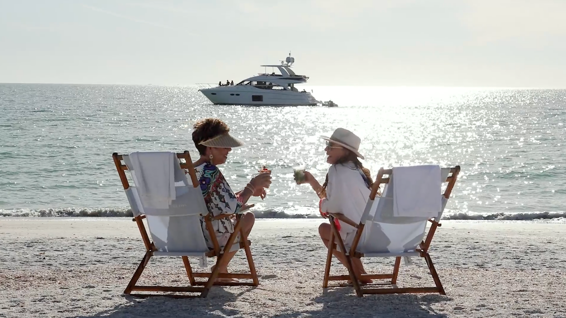 Two people sit in beach chairs facing the sea, chatting, with a yacht in the background.