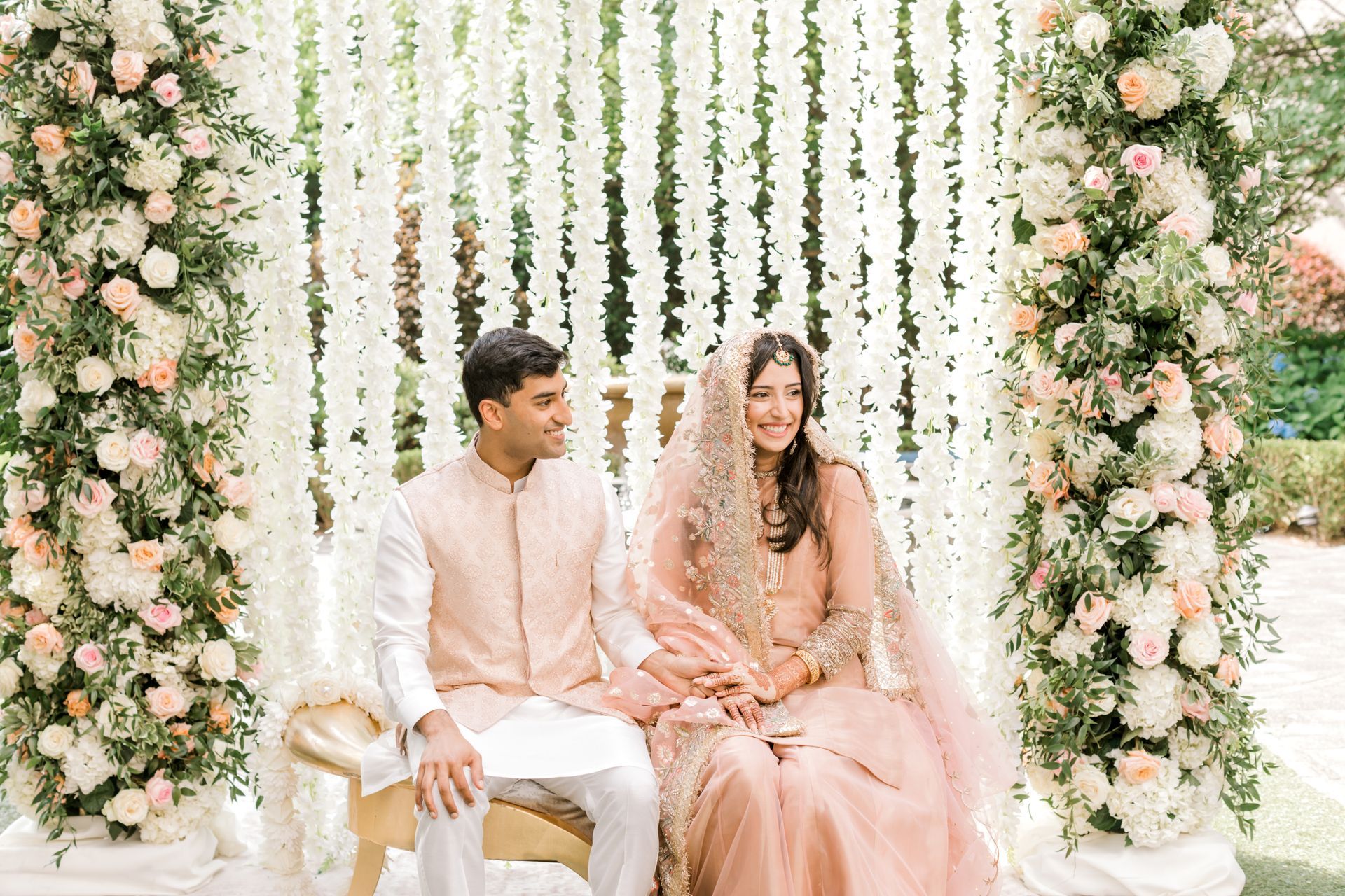 A bride and groom are sitting in front of a wall of flowers.
