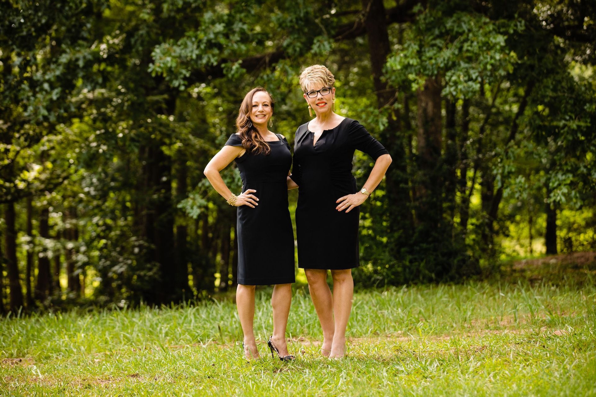 Two women in black dresses pose outdoors in a grassy area with trees.