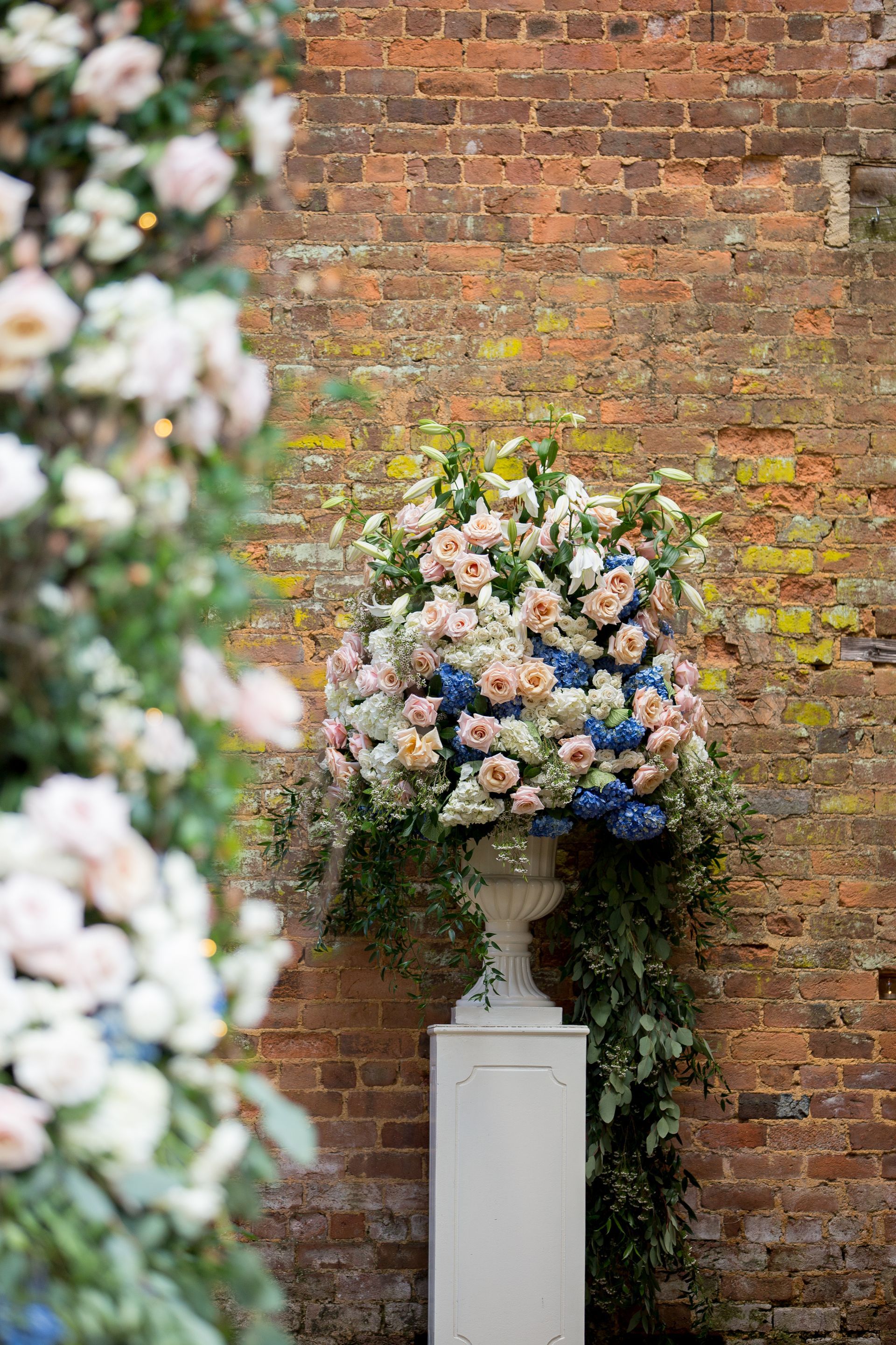 A large vase filled with flowers is sitting on a white pedestal in front of a brick wall.
