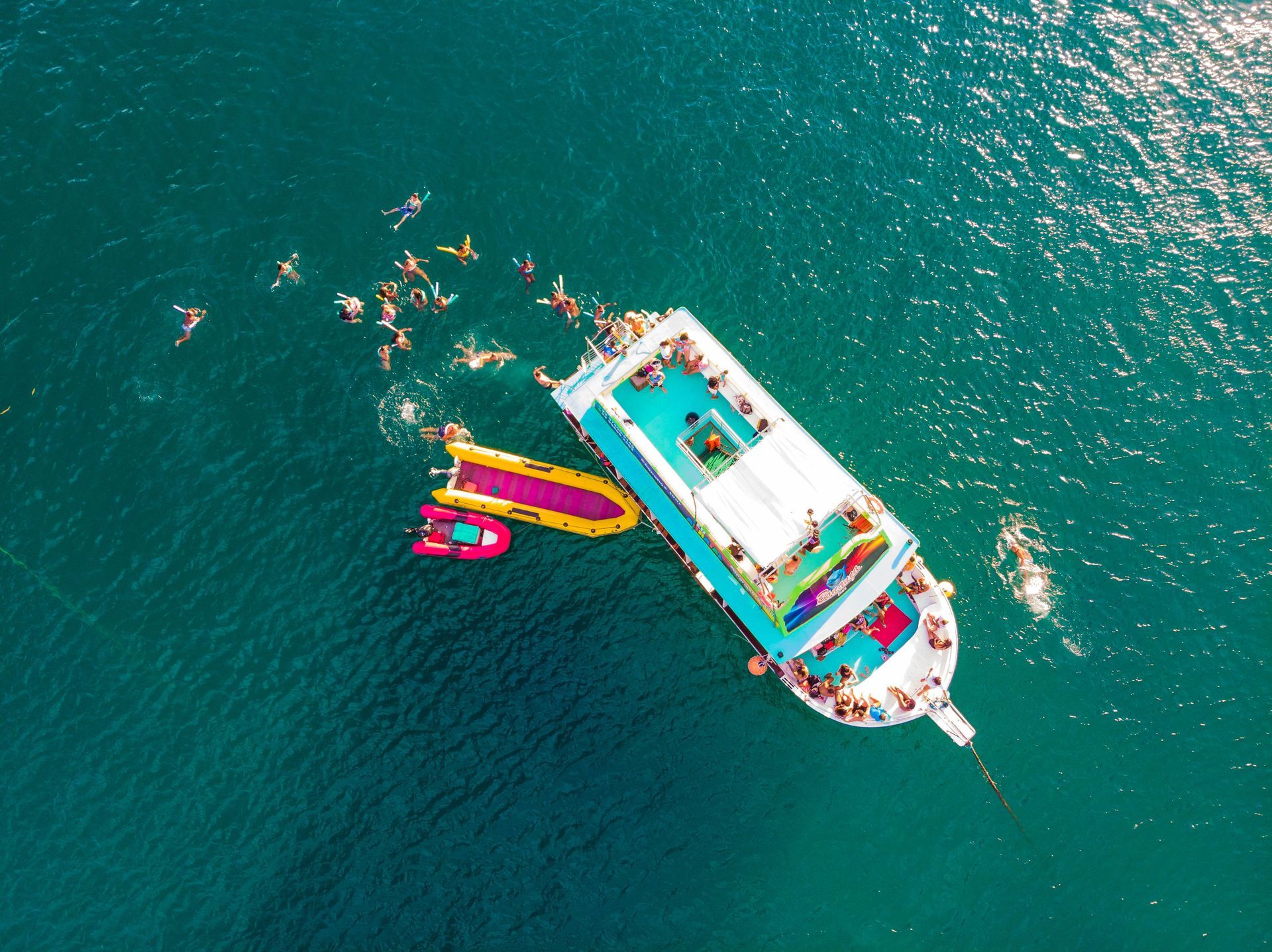 Aerial view of a boat on turquoise water with people swimming and bright pink and yellow inflatables nearby