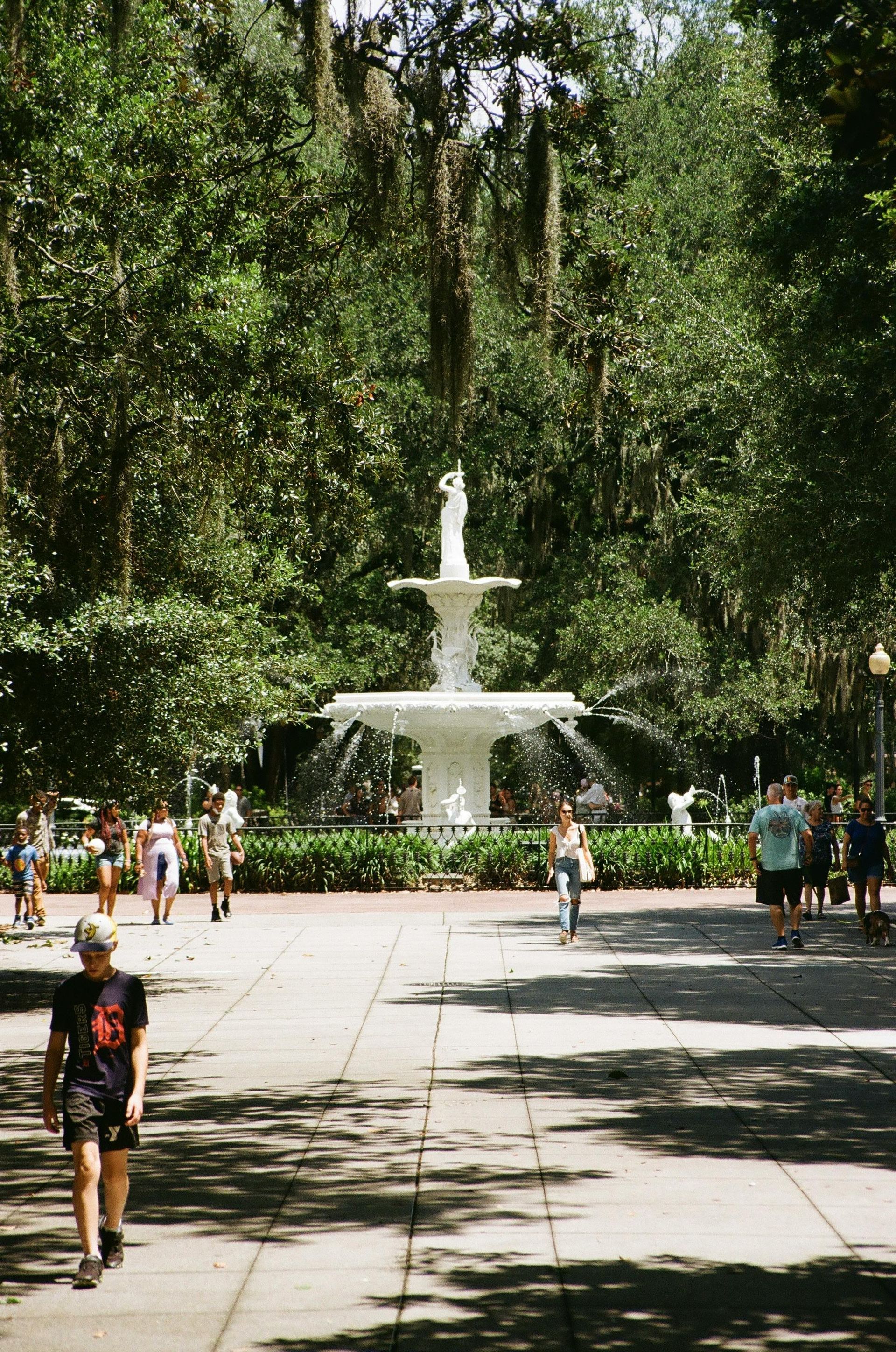Tree-lined park walkway leading to a white fountain, with people strolling in sunlight.