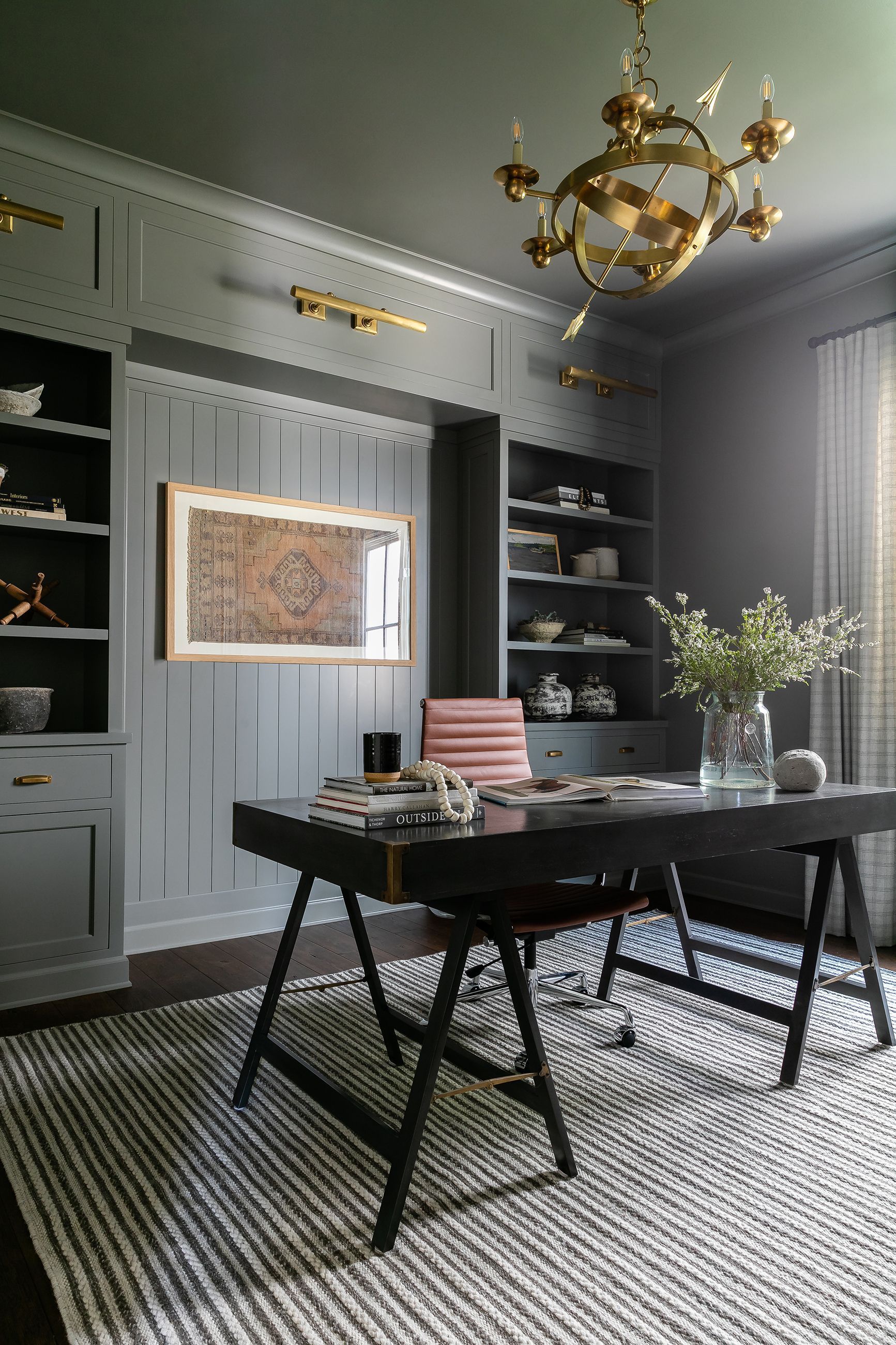 Elegant home office with built-in bookshelves, dark desk, and ornate chandelier. Dark gray walls, light rug.
