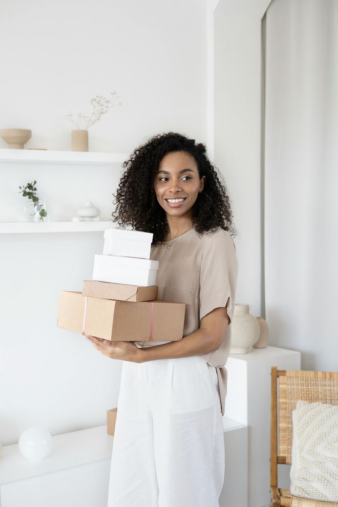 Woman smiling, holding stack of boxes indoors, white walls, neutral clothing.