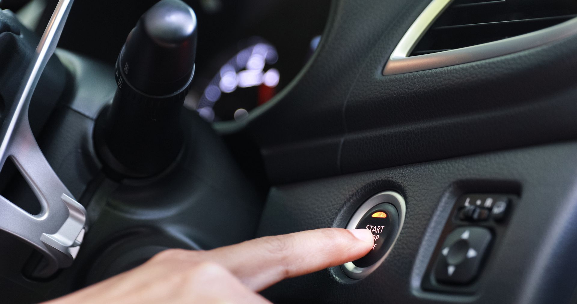 Finger pressing ignition button on a car dashboard near the steering wheel