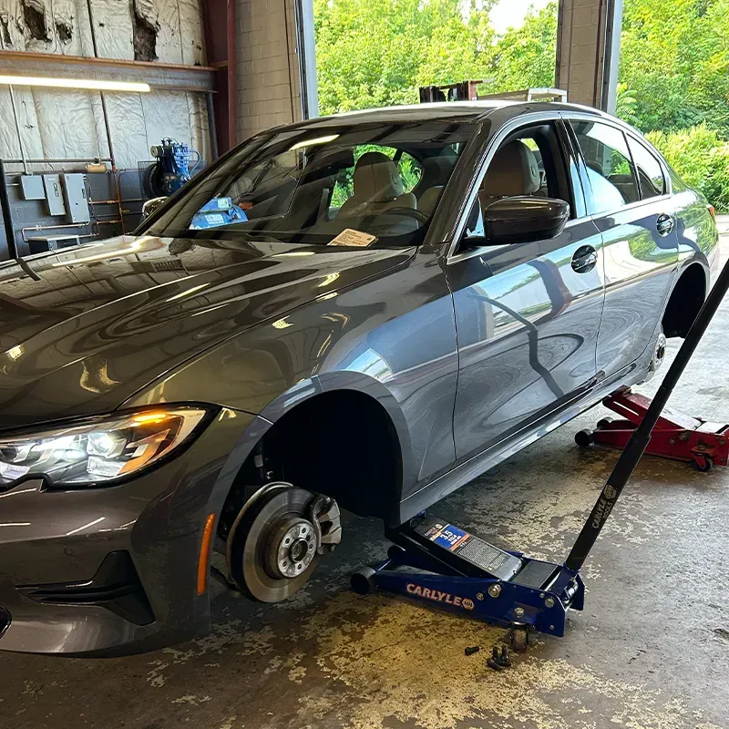Silver car lifted on jack in garage with front wheel removed and brake exposed