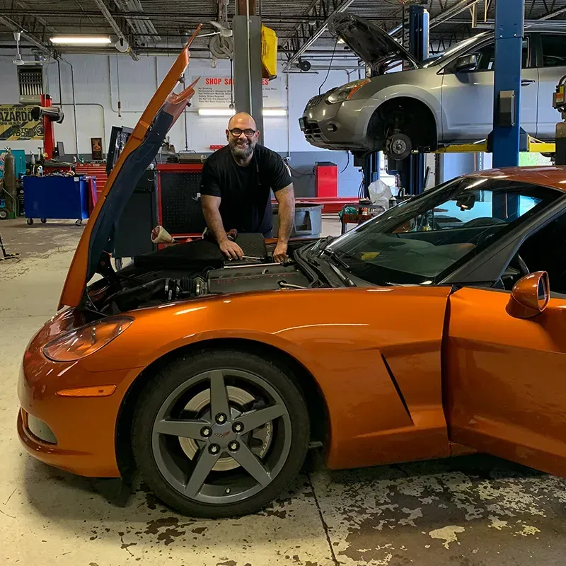 Orange sports car in a repair shop with its hood open, mechanic standing beside it, and another car lifted in the background