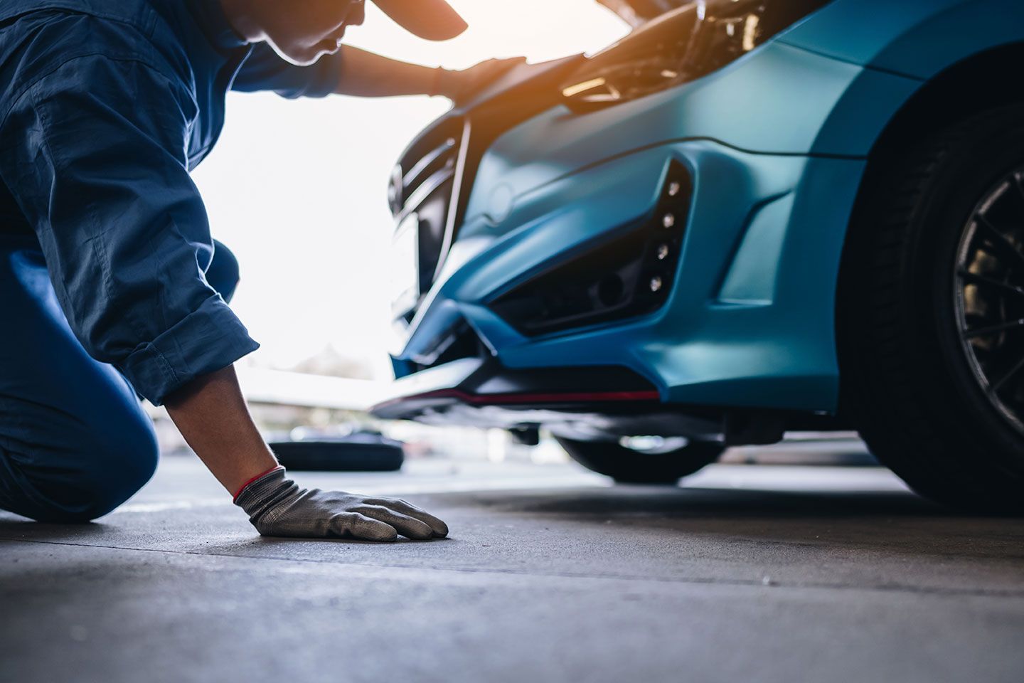 Mechanic kneeling beside a blue sports car, inspecting the front bumper in a garage.