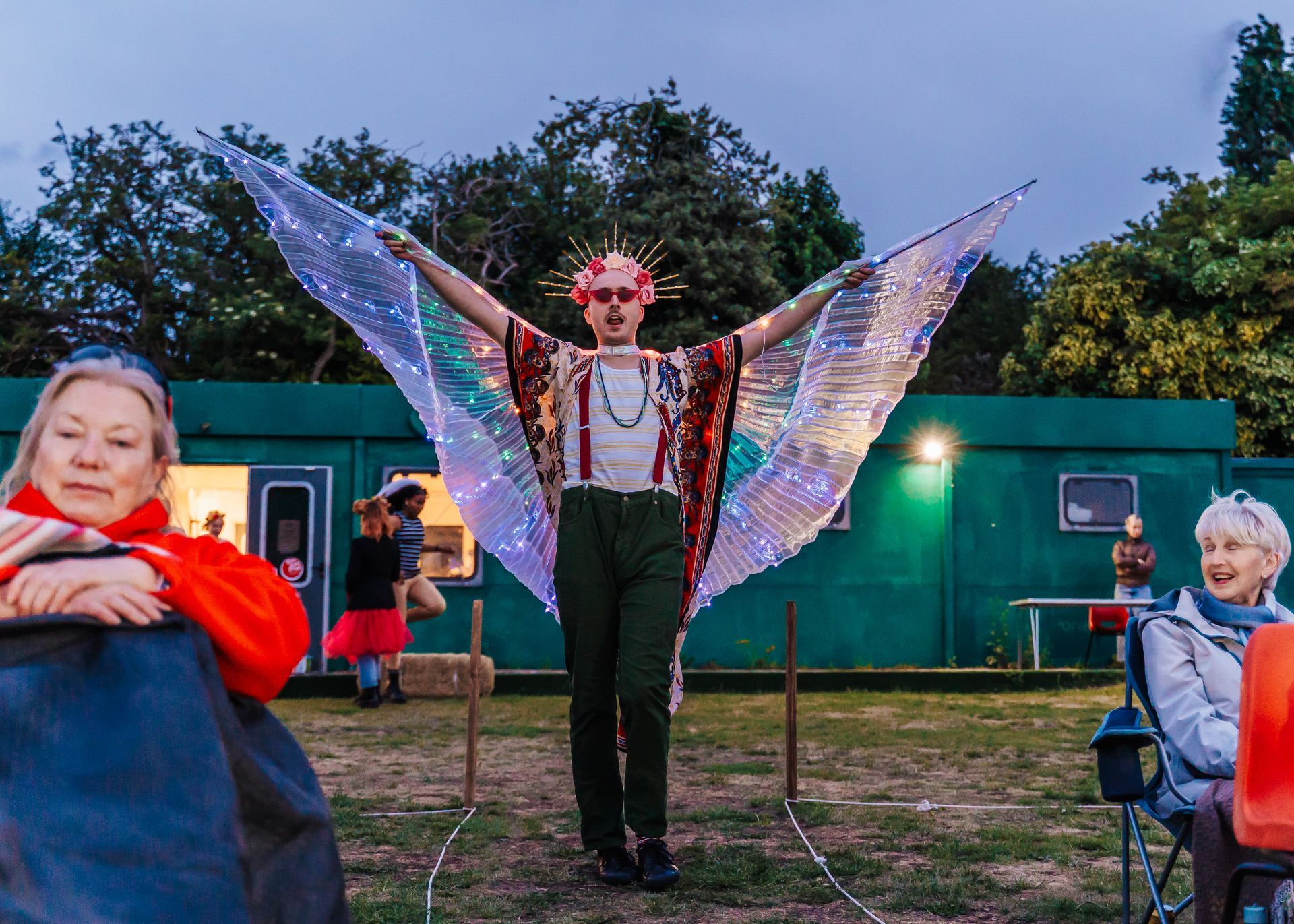 Person in sparkling wing costume standing outdoors with arms raised at an evening event