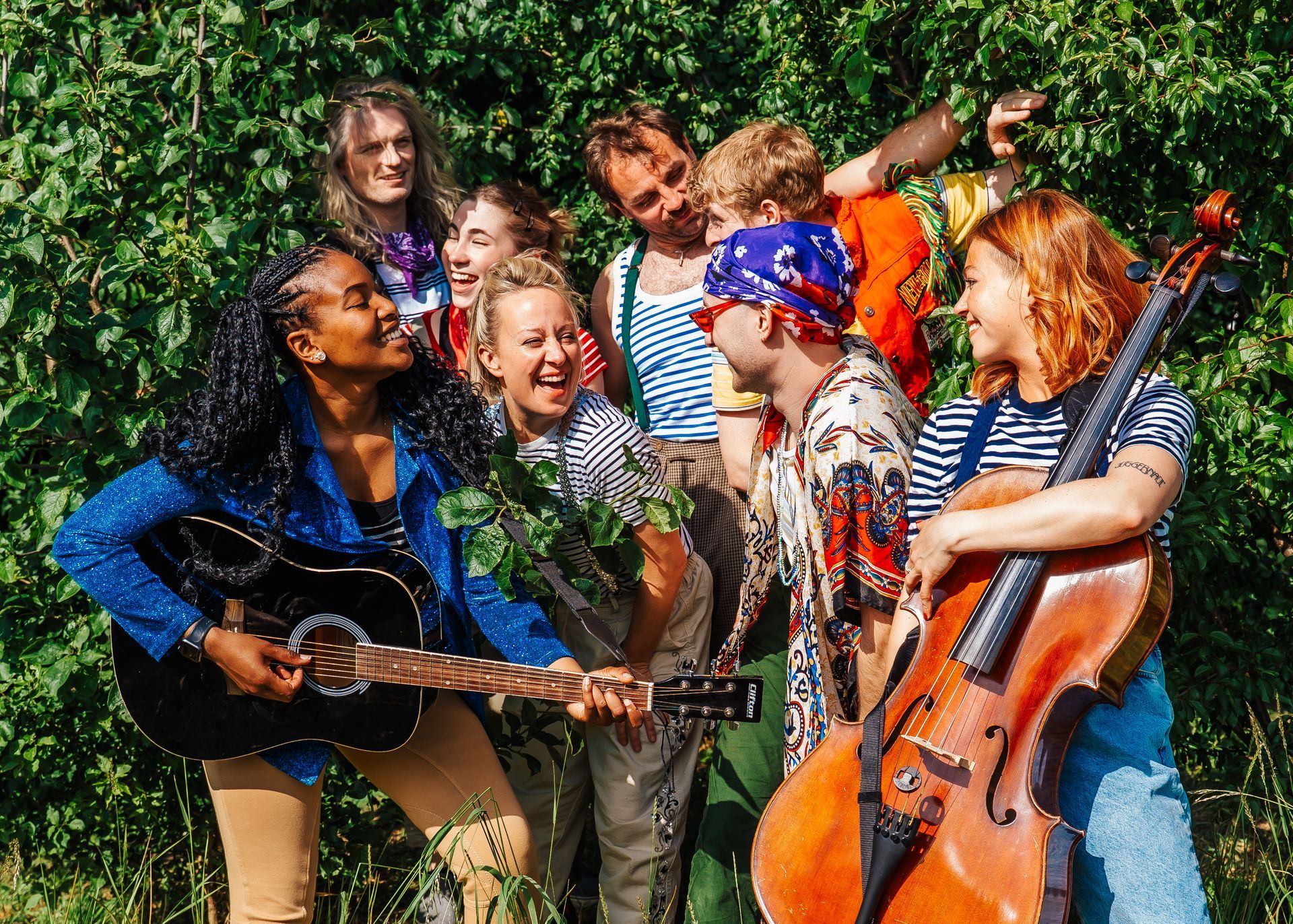 Group of musicians outdoors, smiling and playing guitars and a cello in front of green foliage