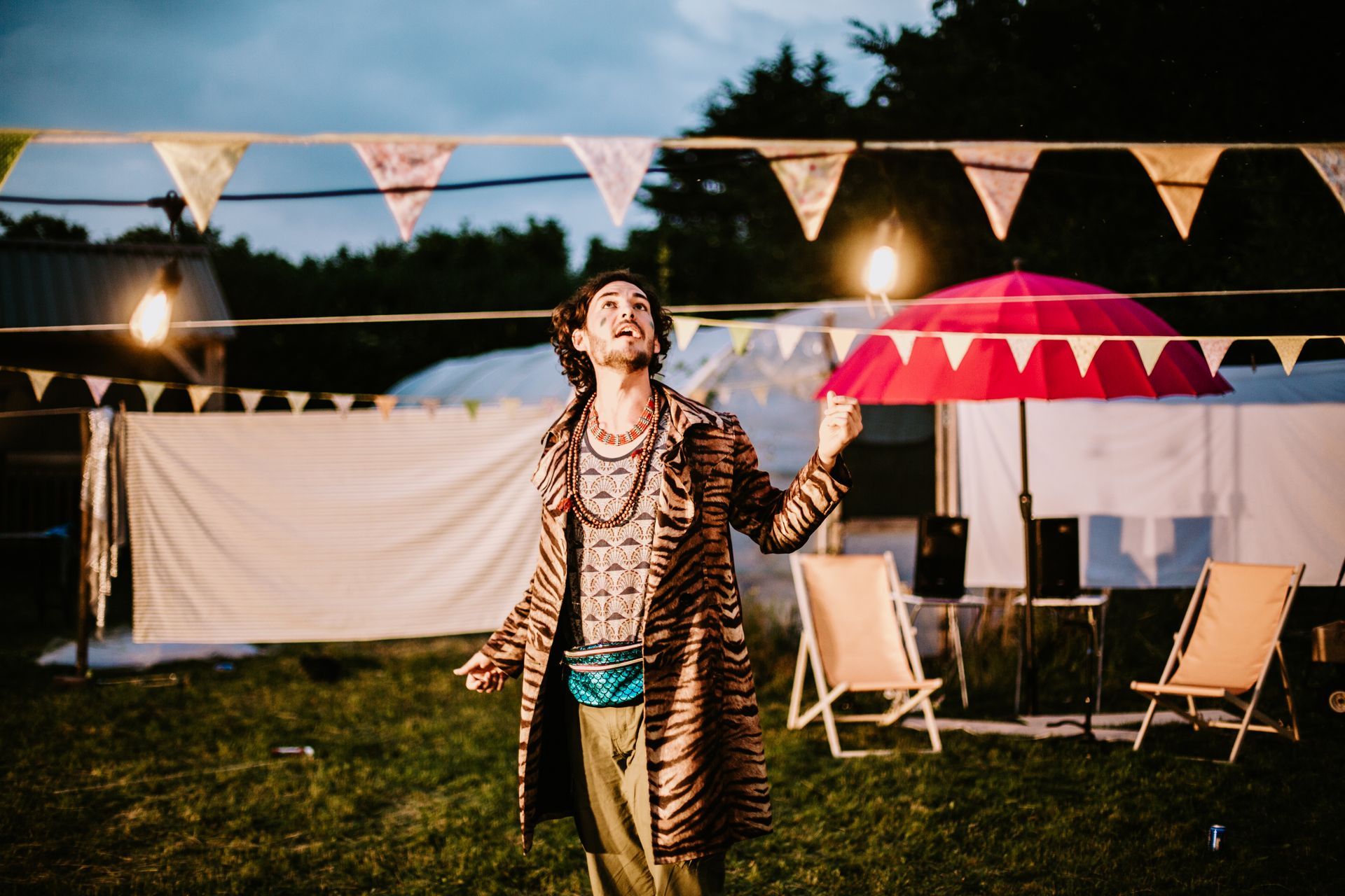 Person in tiger-print coat holding a red umbrella at an outdoor evening party with bunting and chairs.
