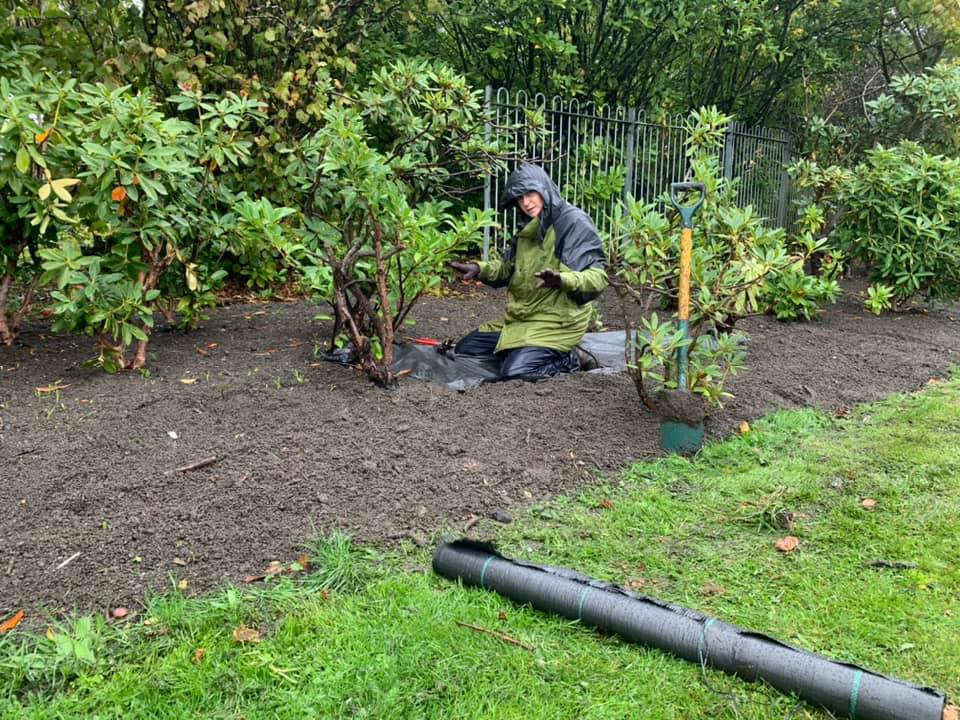 A man in a green jacket is kneeling on the ground in a garden.