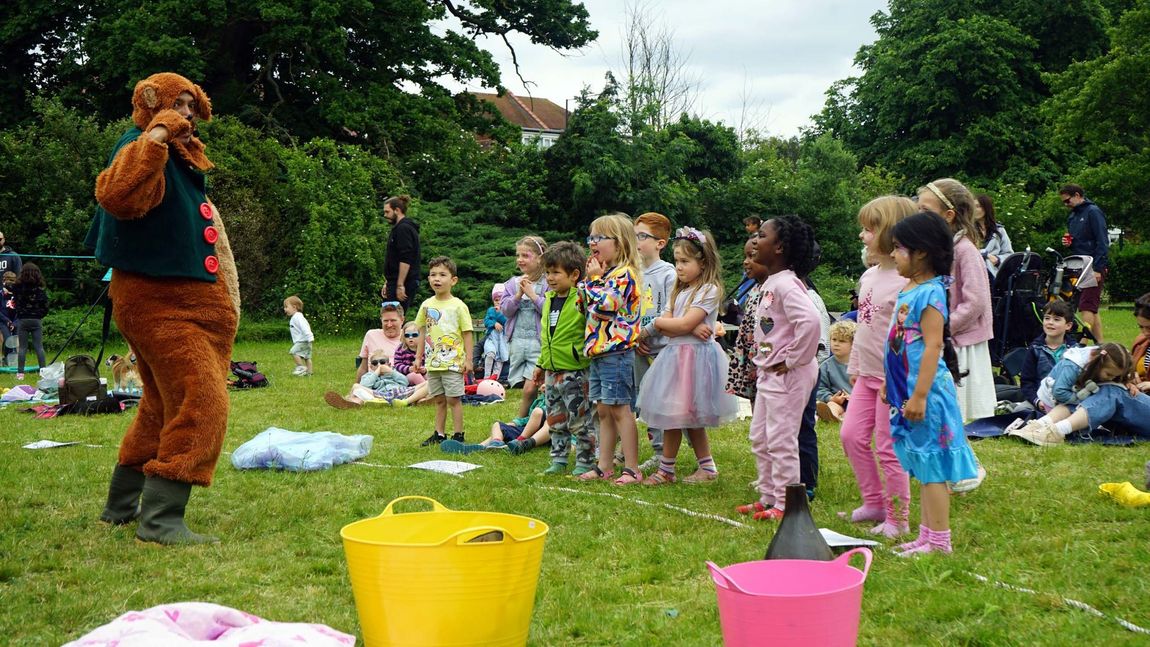 Children watch a person in a brown costume entertain them at an outdoor park party with colorful buckets nearby