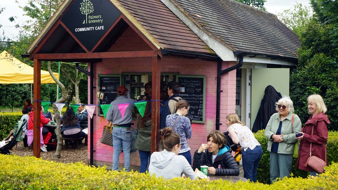 People gathered outside a small red kiosk under a tree, chatting and sitting on benches.