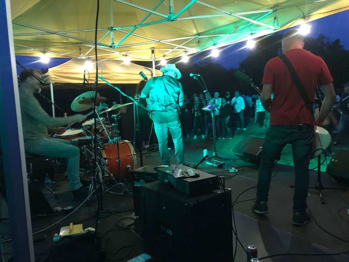 Live band performing on a small stage under green and yellow lights, with drums, guitar, and audience visible.