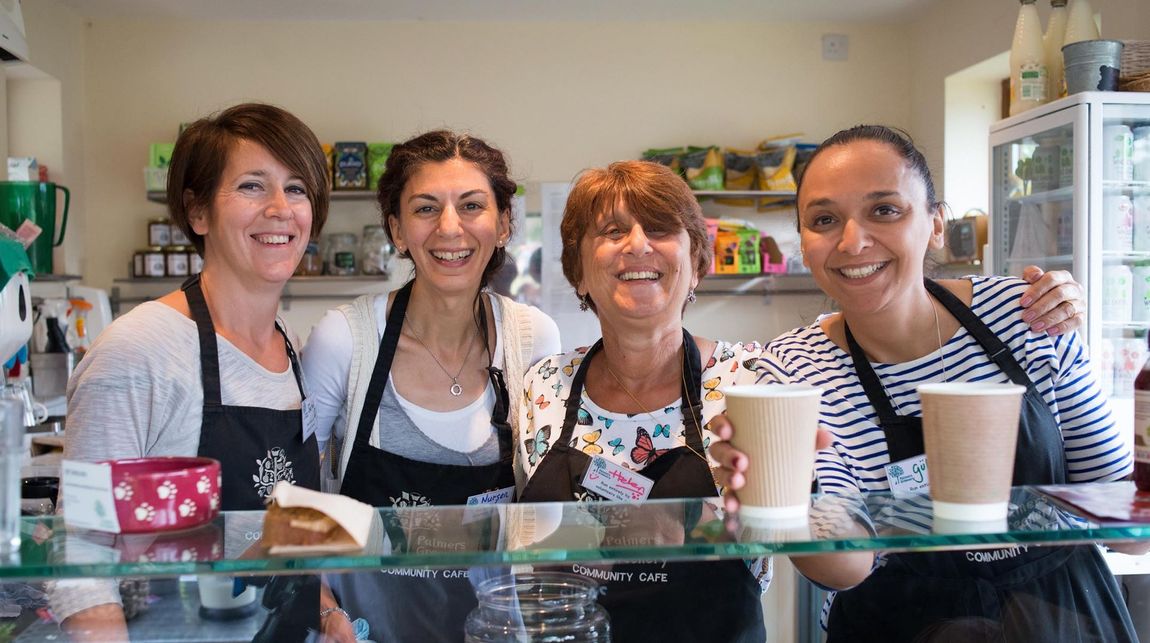 Four people smile behind a café counter with drinks and baked goods displayed in front.
