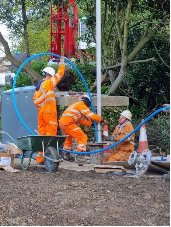 Construction workers in orange safety gear installing pipes beside a red drilling rig.