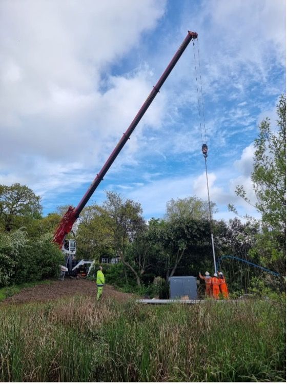 Crane hoisting equipment at a wetland site under a cloudy sky, with workers below.
