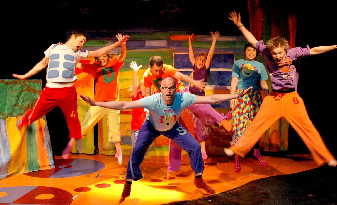 Seven dancers in bright costumes pose mid-performance under colorful stage lights.