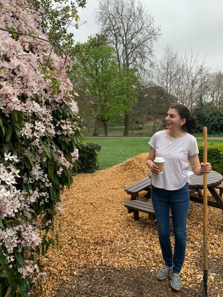 A woman is standing in a park holding a broom and a cup of coffee.